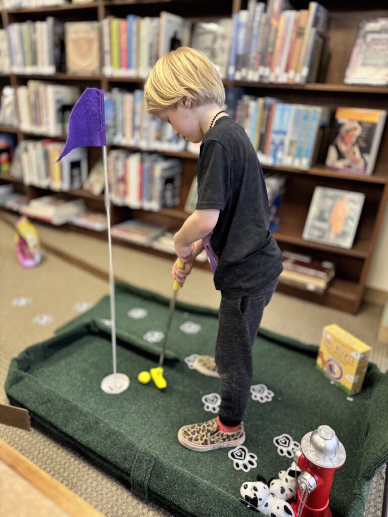 A young child putting a ball into a flagged hole with library book shelves in background