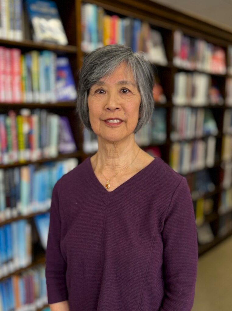 Portrait of Debra DeMeis wearing purple shirt standing in front of book stacks