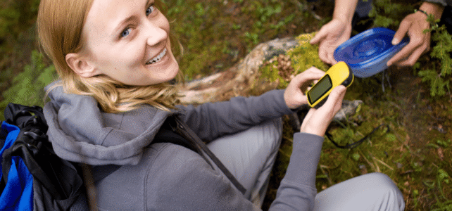 Sewickley Valley Geo-Trail with Fern Hollow A smiling person holding a yellow GPS device and a blue container while sitting in the woods.