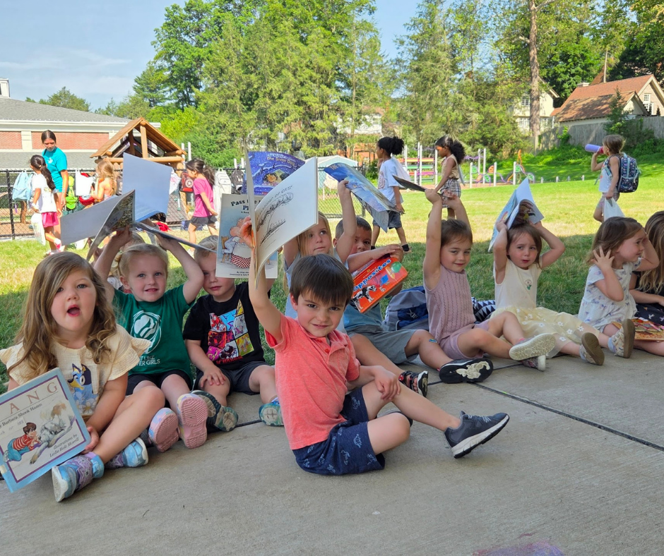 A group of young children sit on the ground outdoors, holding up open books.