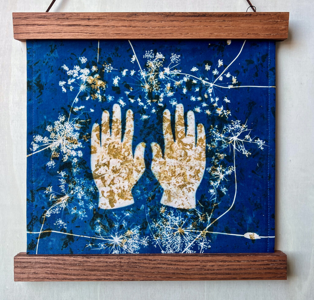 Cyanotype print of hands and botanicals on dark blue fabric with wooden frame on top and bottom