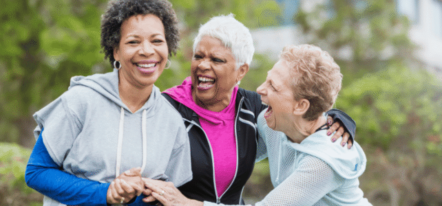 Three people smiling and hugging outdoors in a park, with one person standing in front and two others leaning in affectionately as they embrace.