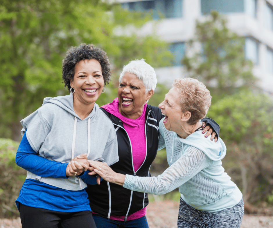 Three people smiling and hugging outdoors in a park, with one person standing in front and two others leaning in affectionately as they embrace.