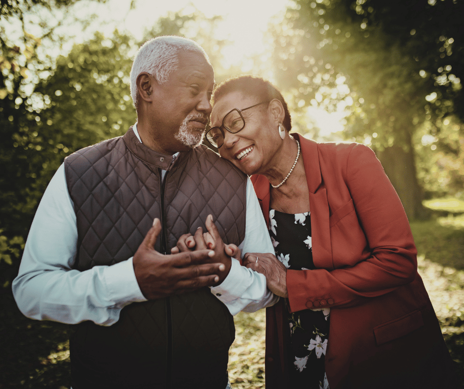 An older couple stands in a sunny park, smiling and holding hands as one gently embraces the other, with trees glowing in golden light.
